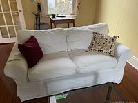 Full view of white Ikea loveseat with burgundy and multi-color throw pillows, placed on wood floor in living room.