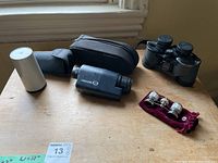 Overview photo showing all four items on a wooden table near a window including opera glasses, Bushnell binoculars, Night Owl Optics monocular, and a compact monocular.