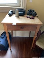 Front view of vintage wooden side table with items on top including binoculars and other small items, showing drawer and turned legs.