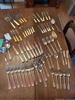 Full overview of all flatware including forks, knives, spoons, and serving utensils laid out on table showing variety of handle styles and sizes.