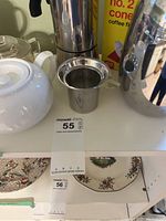 Close-up photo of white ceramic teapot, stainless steel espresso pot, tea strainer, and stainless steel percolator on shelf.