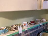 Seven teacups and saucers arranged in a row on a kitchen counter showing a variety of colors and patterns including turquoise, black, floral, and red patterns.