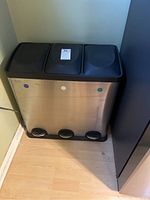 Front view of the stainless steel pedal bin showing three compartments with black lids and three foot pedals below, placed on a wooden floor next to a wall and a piece of furniture.