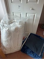 Twin-sized white duvet packaged in clear plastic, standing upright on carpeted floor next to closed white door.