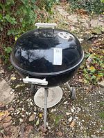 Full view of black Weber kettle charcoal BBQ grill with lid, showing metal legs, wheels, and ash catcher tray beneath
