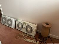 Sunbeam and Honeywell window air conditioners side by side on carpet against wall with Lasko space heater and power strip on floor