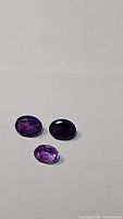 Three oval cut amethyst gemstones arranged on a neutral background, showing color and clarity variations.