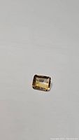 Photo of the natural golden citrine emerald cut gemstone on a white surface, showing color and facet details.