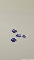 Four pear-shaped natural blue-purple tanzanite cabochons lying flat on a light surface, showing color and size differences with some visible inclusions.