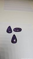 Three polished natural charoite gemstones in deep purple with black and white swirls on a white background.