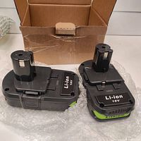 Two 18V lithium ion batteries with labeling showing Li-ion 18V, placed on bubble wrap with an open cardboard box behind them.