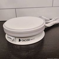 Side and angled view of a white motorized rotating turntable showing the power port and GLO3D branding, model Stand-313, with attached white power cord.