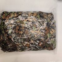 Ghillie suit folded and sealed in clear plastic packaging showing full camo pattern.