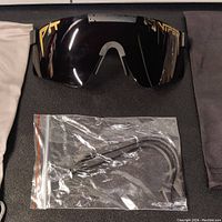 Front view of black Pit Viper sunglasses with reflective black polarized lens, flanked by two fabric pouches and a small bag with extra ear hooks inside.