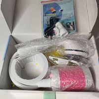 Top view of the unboxed camping shower components including shower head, hose, suction cup base, and charging cable all wrapped in bubble wrap inside the box.