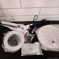 Parts of the RBG LED magnifying desk lamp including the circular LED light ring, adjustable arm with spring, remote control, power cord, weighted lamp base, and desk clamp, all wrapped in protective plastic.