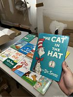 Photo showing most of the Dr. Seuss books arranged on a table including The Cat in the Hat cover held by hand.