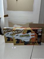 Two boxes of Nerf Ultra AMP blaster toys on a table, showing front of packaging that highlights brand, model, and firing distance.