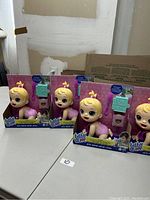 Four brand new Baby Alive dolls in original packaging, each with blonde hair in a small ponytail and pink outfits, sitting on a white surface with a partially visible cardboard box behind them.