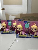 Four boxed Baby Alive dolls standing side by side, showing front of packaging and dolls with blonde hair and pink outfits.