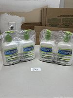 Photo showing two shrink-wrapped pairs of Cetaphil lotion bottles on a table in front of cardboard boxes.