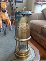 Front view of brass Davy lamp showing 'Cymru' badge with dragon emblem and partial corrosion on top.