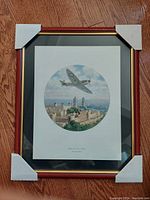 Framed wood picture of 'Spitfire Over Tower Bridge' by John Young wrapped with corner protectors on hardwood floor.