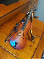 Photo showing the miniature wooden cello with the bow resting on a wooden surface and a brass stand supporting the cello, highlighting craftsmanship and vintage condition.