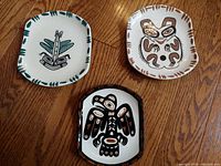 Three vintage Lambert Potteries Ltd. hand-painted pottery plates on hardwood floor. Plates feature West Coast Indian designs in black, brown, orange, and green.