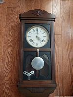 Front view of vintage mechanical wall clock with pendulum and winding key, sitting on wooden floor