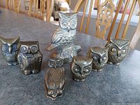 Seven brass owl figurines arranged on table showing a view from front and side. Different sizes and shapes, the tallest owl stands on a branch base.