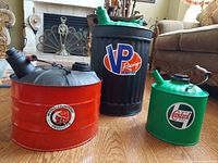 Set of three custom vintage painted fuel cans on wooden floor near furniture. Showing red McColl-Frontenac, tall black VP Racing, and green Castrol cans with spouts and handles.