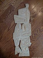 Top-down photo of an unfinished wood carving featuring a bear holding a salmon in a First Nations art style. Carving is laid on a wooden floor.