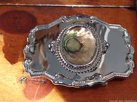 Top angle of silver plated belt buckle showing the ornate lifted edges and oval mother of pearl central inlay with beaded border.