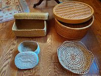 Four handcrafted indigenous baskets arranged on a wood floor, showing boxy rectangular basket with lid, round wooden steamer-style basket, shallow six-point basket, and oval basket with braided handle.
