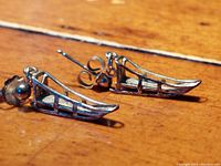 Close-up photo of the pair of silver dog sled shaped earrings lying on a wooden surface showing details and structure.