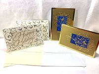 Four large embossed Christmas cards with envelopes. Two gold cards with embossed tree design over blue background, two cream cards with embossed 'Happy Holidays' text and golden swirls. All accompanied by envelopes.