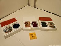 Three vintage porcelain music boxes in molded foam packaging with red lids. Each box comes with its own container and Certificate of Authenticity papers are visible for two boxes.