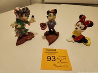 Three Disney figurines: Mickey in purple, Minnie in green, and separate ceramic Minnie on a white surface with auction label