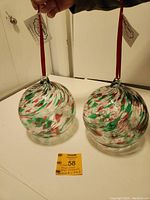 Pair of large handmade blown glass globes with red, green, and white swirls hanging from red ribbons, placed on a white surface with tags visible.