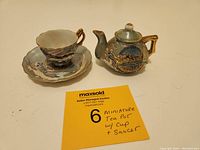 Miniature tea cup, saucer, and tea pot placed on flat surface with auction card. Shows overall size and Mt Rushmore decoration.