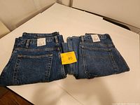 Two pairs of folded blue jeans laid side by side on white surface with yellow lot number card.