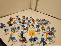 Photo showing entire lot of 32 Smurf PVC figures arranged on white surface with yellow note