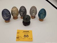 Front view of 5 egg-shaped carved stones with etched designs and 1 green stone sphere on a carved wooden stand. Stones vary in color, including purple, beige, dark gray, pink, and blue-green.