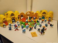 Wide view of three yellow plastic Smurf school playset buildings and 32 assorted blue PVC Smurf figures arranged in front. Includes interactive green base with buttons.