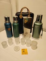 All items grouped together: three vintage Stanley thermoses, metal box with frosted plastic stackable cups, and navy blue carrying bag with leather handles.
