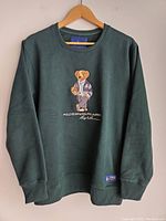 Front view of dark green crewneck sweatshirt hung on wooden hanger against white background, showing overall design, fit and condition.