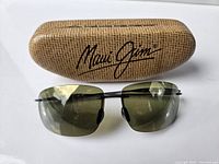 Front view of Maui Jim sunglasses with green lenses and textured Maui Jim case in the background.
