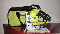Ryobi biscuit joiner displayed alongside green and black Ryobi branded carrying bag and instruction manual, placed on wooden surface against white background.