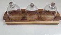 Front view of mango wood serving board holding three clear crystal cloche lids, showing size and fit of lids on board.